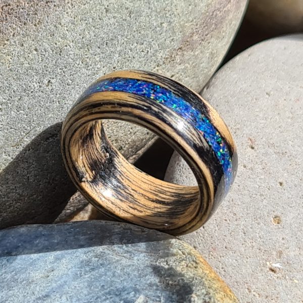 Hand crafted ebonised oak ring with blue opal inlay resting on beach stones.