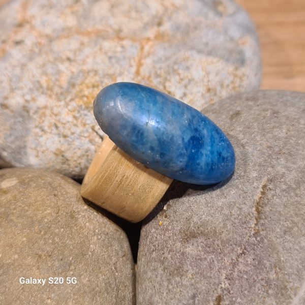 A neon Apatite Oval Cabochon on an ash bentwood ring. It is resting on some beach stones.